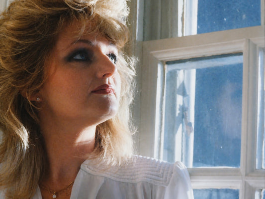 Blonde woman in a white blouse gazing upwards beside a blue-lit window.
