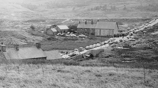 Residents and vehicles moving through Capel Celyn in October 1963, with stone buildings beside the village road