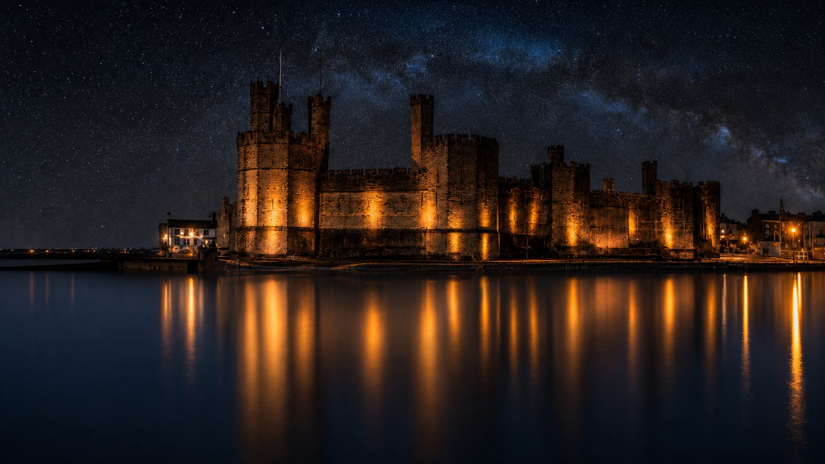 Caernarfon Castle illuminated at night, with golden lights reflected across calm harbour water.